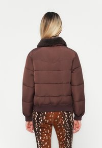 Giacca puffer marrone con un colletto in finta pelliccia testurizzato e polsini a coste, caratterizzata da un design trapuntato e una vestibilità accorciata. Indossata con pantaloni a fantasia.