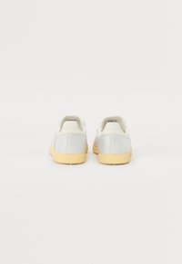 Ett par vita låga sneakers med beige gummisulor, visade bakifrån på en neutral vit bakgrund.