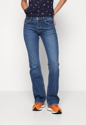 Person trägt blaue Straight-Leg-Jeans, ein dunkles Oberteil mit kleinen Blattmustern und leuchtend orangefarbene Sneakers, steht mit den Händen in den Taschen.