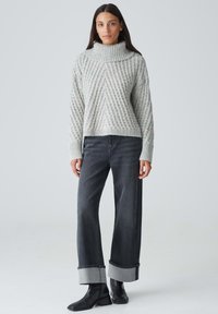 Grauer gestrickter Pullover mit hohem Funnel-Neck, der ein strukturiertes diagonales Muster aufweist; kombiniert mit weit geschnittenen schwarzen Jeans und Stiefeletten.
