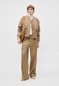 Bež bomber jakna z elastičnimi manšetami, kombinirana s širokimi bež hlačami; model drži rjavo torbico na ramenu; viden bel vodniški pulover.