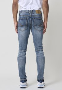 Vaqueros skinny de denim azul con un acabado descolorido, cinco bolsillos y detalles de costura en beige. Presenta una parche de cuero en la parte trasera de la cintura.