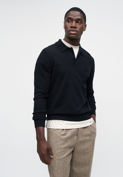 Homem vestindo uma camisa polo de manga longa preta sobre uma blusa branca e calças bege texturizadas, de pé com uma mão no bolso, olhando para o lado.