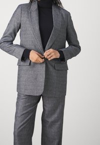 Blazer gris sur mesure en tissu texturé avec revers à encoches, deux poches avant et une fermeture à bouton, assorti d'un pantalon coordonné.