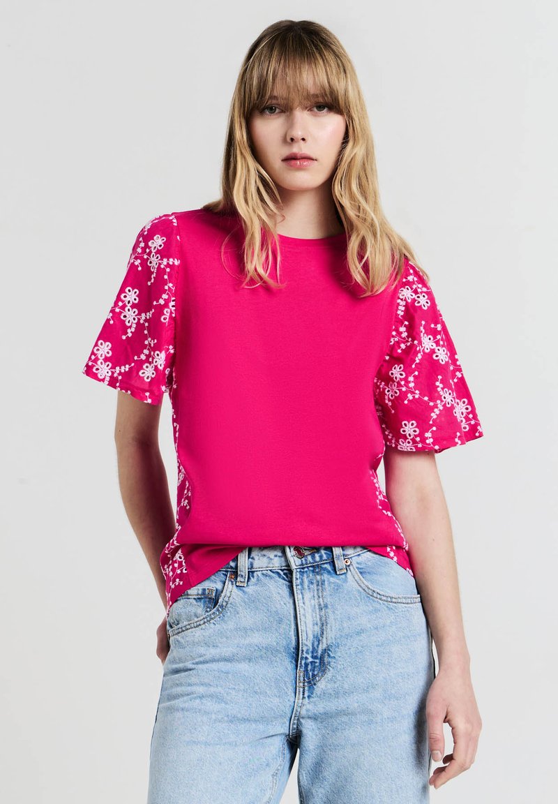 Top fucsia con maniche corte a fantasia floreale, caratterizzato da un taglio dritto e una texture morbida. Abbinato a jeans chiari in denim.