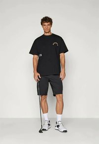 Zwarte oversized t-shirt met oranje tekst en een korte broek in een bijpassende kleur. Het model draagt witte sneakers met zwarte accenten en houdt een golfclub vast.