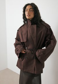 Kvinna med lockigt mörkt hår iklädd en överdimensionerad brun trenchcoat med bälte, stående mot en helt vit vägg.