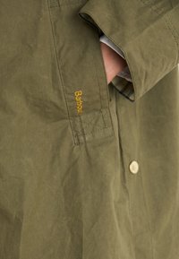 Olivově zelená bunda s kapsou, žlutou výšivkou "Barbour", stříbrným knoflíkem a texturovanou, lehkou tkaninou.