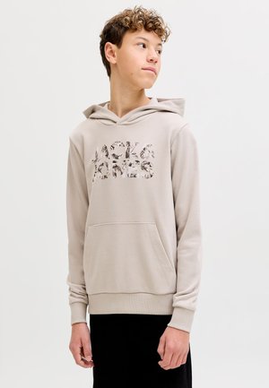 Băiat adolescent cu păr creț, purtând un hanorac bej Jack & Jones, privind în lateral pe un fundal simplu și luminos.