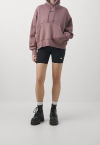 Vaaleanpunainen ylisuurikokoinen huppari, jossa etutasku ja musta logo, yhdistettynä mustiin pyöräilyshortseihin, joissa on pieni logo, sekä mustiin nauhallisiin saappaisiin.