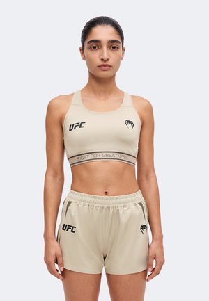 Femme portant un soutien-gorge de sport et un short beige UFC avec le texte "Fight for Greatness", debout, face à l'avant sur un fond uni.
