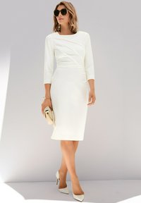 White fitted dress with three-quarter sleeves, textured front detailing, and a knee-length hem. Paired with white pointy heels and a clutch.