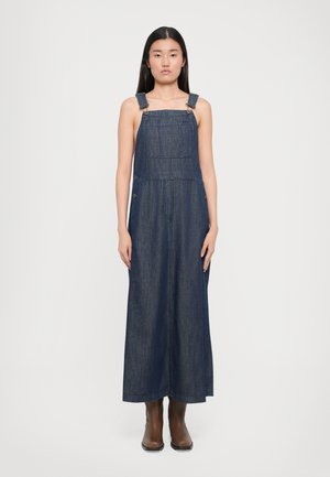 Denim overall jurk in donkerblauw, met verstelbare schouderbanden, voorzakken en een losse pasvorm, gecombineerd met bruine enkellaarsjes.