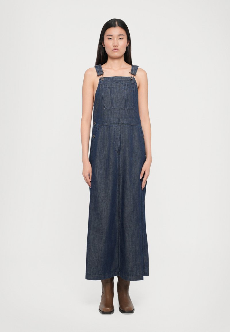 Rochie salopetă din denim de culoare albastru închis, având bretele reglabile, buzunare frontale și o croială lejeră, asortată cu cizme până la gleznă de culoare maro.