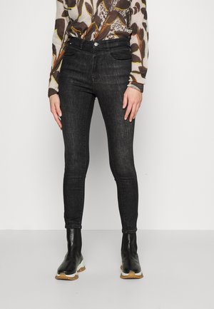 Femme portant un jean skinny noir taille haute, un haut à manches longues à motif, et des bottines noires avec des semelles épaisses blanches.