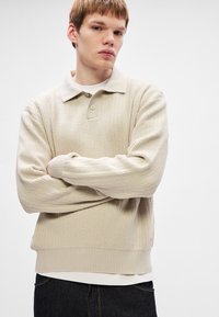 Pulover din tricot bej cu textura rib, cu un guler polo, având o prindere cu trei butoane și manșete rib, purtat peste un top alb.