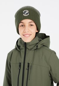 Berretto lavorato a maglia verde con patch rotondo, abbinato a una giacca verde scuro con tasche con zip e colletto alto.