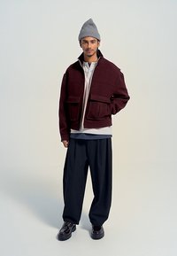 Burgundská bomber bunda se dvěma předními kapsami, kombinovaná se světle šedým topem a volnými černými kalhotami. Doplňky zahrnují šedou čepici.