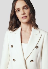 Dwurzędowa biała marynarka wykonana z gładkiego materiału, ozdobiona ciemnoskalnymi guzikami i ostrymi klapami, o dopasowanym kroju.