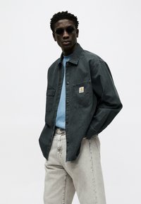 Férfi sötétzöld gombos ingben, kék póló felett, világosszürke farmerben és fekete kerek napszemüvegben, egyszerű világos háttér előtt áll.