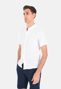 Camicia bianca a maniche corte con bottoni, texture in lino con colletto alla francese e vestibilità rilassata, abbinata a pantaloni navy. Design semplice, senza fantasie.