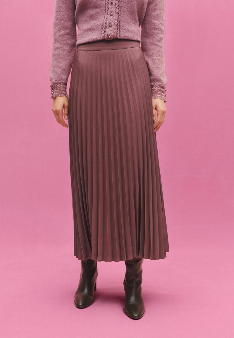 Femme portant un pull mauve à manches longues avec des poignets en dentelle, une jupe plissée violette taille haute, et des bottines noires sur un fond rose.