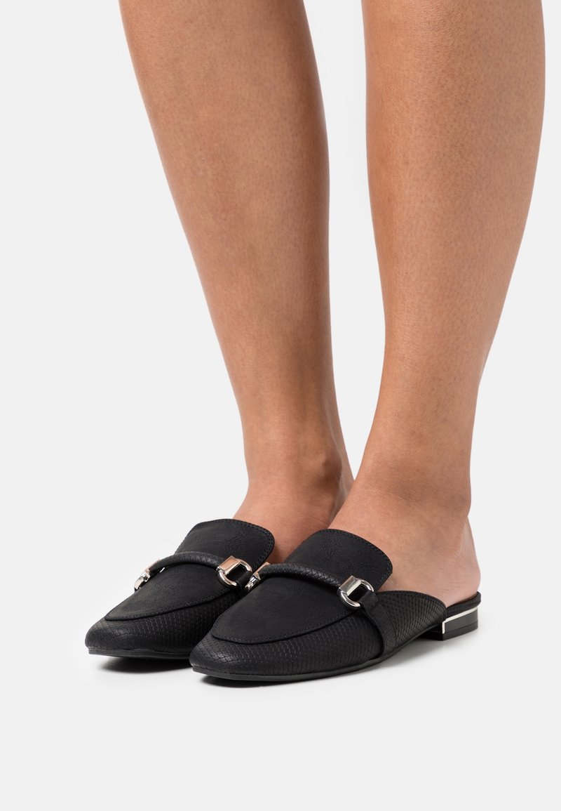 Des chaussures noires à enfiler avec une surface texturée, présentant un bout arrondi, une sangle décorative avec des accessoires en argent, et un petit talon carré.