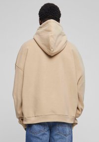 Beige, oversized hoodie met een grote capuchon, zachte textuur en verlaagde schouders. Gecombineerd met blauwe denim jeans die aan de onderkant zichtbaar zijn.