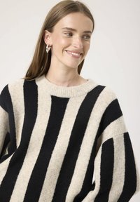 Frau lächelt, trägt silberne Creolen und einen beigen Pullover mit breiten senkrechten schwarzen Streifen, vor einem schlichten hellen Hintergrund.