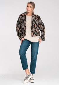 Braune und schwarze, diamantgefleckte Kurzjacke mit großen Revers, kombiniert mit einem beigen Strickpullover und blauen Jeans. Die Sneaker haben ein Leopardenmuster.
