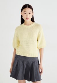 Suéter de malha amarelo claro de mangas curtas, com decote redondo e bainha canelada, combinado com uma saia plissada cinza-escuro.