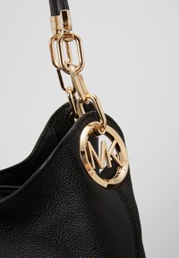 Coin de sac à main en cuir texturé noir avec une bandoulière chaîne dorée et un charm rond avec le logo MK attaché près du bord supérieur.