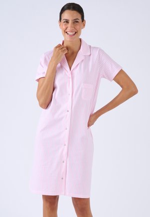 Vrouw glimlacht, draagt een roze-wit gestreepte knoopjurk met korte mouwen tot de knie, staat met één hand op de heup.