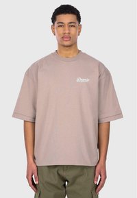 Übergroßes beigefarbenes Baumwoll-T-Shirt mit kurzen Ärmeln, das auf der linken Brustseite den Schriftzug "Dropize from the Archives" in weißer Schrift trägt.