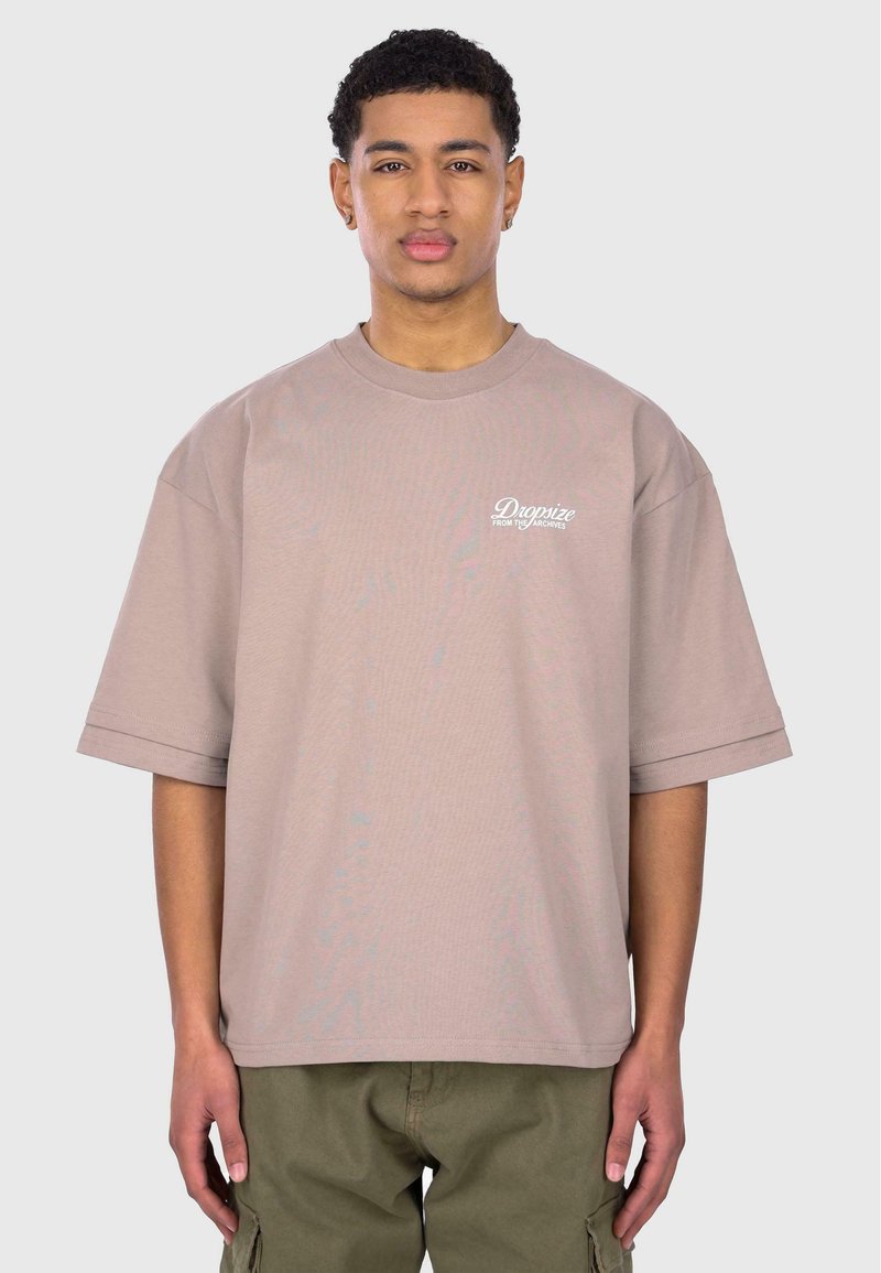 Übergroßes beigefarbenes Baumwoll-T-Shirt mit kurzen Ärmeln, das auf der linken Brustseite den Schriftzug "Dropize from the Archives" in weißer Schrift trägt.