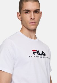 Camiseta de algodón blanca con el logo de "FILA" en bordado negro y rojo y la frase "ESTABLECIDO EN 1911" en texto negro. Mangas cortas.