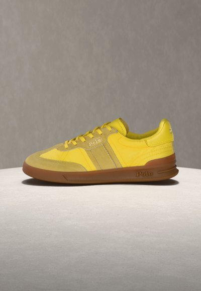 Κίτρινο αθλητικό παπούτσι Polo με μπεζ σουέντ λεπτομέρειες και καφέ σόλα, τοποθετημένο σε στρογγυλή επιφάνεια ανοιχτού γκρι χρώματος πάνω σε απλό γκρι φόντο.