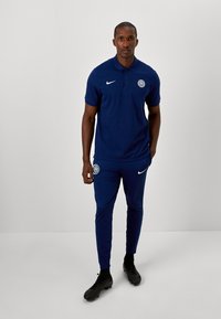 Marineblå polo og matchende bukser lavet af glat stof, med et Nike-logo og klubemblemet, samt sorte sportssko.