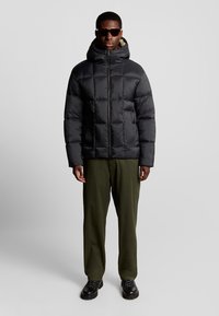 Schwarze Steppjacke mit Kapuze, ausgestattet mit einem gesteppte Design und Reißverschluss. Kombiniert mit olivgrünen Hosen und schwarzen Stiefeln.