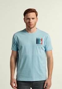 Camiseta de algodón azul claro con un colorido estampado gráfico "Polo" y el texto "valecuatro" en el área del lado izquierdo del pecho. Corte regular, mangas cortas.