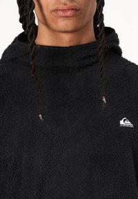 Pullover in pile nero con superficie testurizzata, dotato di collo alto e patch logo laterale. In primo piano è visibile una distintiva acconciatura intrecciata.
