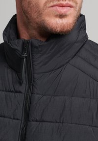 Schwarze Pufferjacke mit hohem Kragen, ausgestattet mit einem Reißverschluss und gestepptem Muster. Das Material wirkt leicht und leicht glänzend.