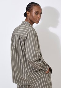 Camisa de manga larga a rayas en verde oliva y crema, con cuello mandarín, corte holgado y bolsillos laterales, hecha de una tela ligera.