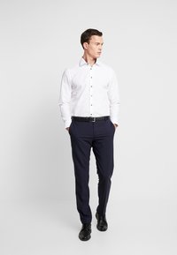 Camicia bianca con bottoni scuri, abbinata a pantaloni eleganti blu navy. Il modello sta in piedi con le mani nelle tasche, indossando scarpe nere e una cintura.
