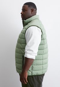 Hellgrüne Steppweste mit horizontalem Quilted-Design. Verfügt über einen hohen Kragen, Reißverschluss-Seitentaschen und einen kontrastierenden Saum entlang der Kanten.