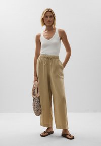 White sleeveless top, light beige wide-leg pants with an adjustable drawstring, and a woven tote bag. Brown flip-flops complete the look.