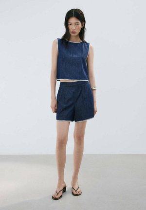 Femme debout portant un crop top en denim bleu foncé sans manches et un short assorti avec un bord blanc, associé à des sandales à talons noires et un bracelet argenté.