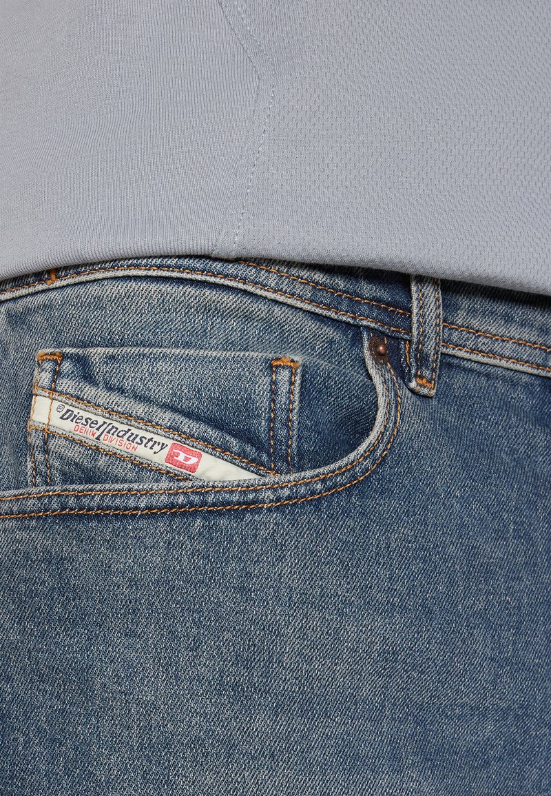 Lichtblauw T-shirt overlapt donkerblauwe denim jeans. De zak toont een wit label met rode accenten en merkdetails. Denimtextuur zichtbaar.