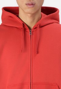 Rote Zip-Hoodie aus weichem Stoff, ausgestattet mit einem Frontreißverschluss, Kordeln und einer gefütterten Kapuze. Glatte Textur ohne sichtbare Muster.