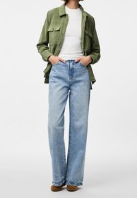 Zielona kurtka sztruksowa z dwoma kieszonkami na piersi, połączona z jasnoniebieskimi, wysokim stanem jeansami i brązowym obuwiem. Pod spodem prosta biała koszulka.